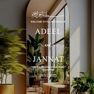 Puede incluir: Un espejo grande y arqueado con marco dorado. El espejo refleja una habitación con plantas y muebles. Texto blanco en el espejo que dice "Bienvenida a la boda de Adeel y Jannat" y una fecha. Decoración de boda.