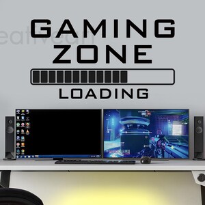 May include: Black and white vinyl wall decal with the text "GAMING ZONE" and a progress bar that reads "LOADING". The decal is displayed on a gray wall above a desk with two computer monitors, a keyboard, a mouse, and speakers.