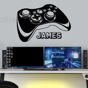 Könnte beinhalten: Schwarz-weiße Silhouette eines Videospiel-Controllers mit dem Namen "JAMES" darunter. Der Controller befindet sich an einer grauen Wand. Im Hintergrund sind zwei Computermonitore und ein Schreibtisch mit Tastatur und Maus zu sehen.