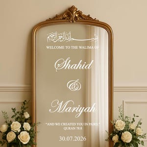 Puede incluir: Un espejo con marco dorado y detalles ornamentales. La superficie del espejo presenta caligrafía y texto en blanco, incluyendo nombres y una fecha. Arreglos florales flanquean el espejo, agregando un toque decorativo al anuncio de boda o evento.
