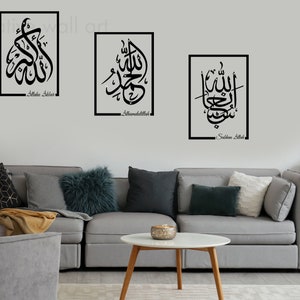 Puede incluir: Tres impresiones de arte de pared enmarcadas en negro con caligrafía árabe en blanco. Las impresiones están colgadas en una pared blanca sobre un sofá gris con almohadas. El texto en las impresiones dice "Allah Subhana Allah", "Alhamdulillah" y "Subhan Allah".