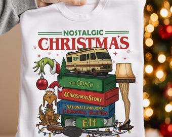 Puede incluir: Sudadera blanca con la frase "NOSTALGIC CHRISTMAS" e ilustraciones navideñas. Incluye una mano del Grinch, una bola roja, una autocaravana y una lámpara de pierna. El diseño también presenta una pila de cintas VHS.