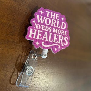 Può includere: Un portabadge con un design glitterato rosa. Il portabadge presenta il testo bianco "THE WORLD NEEDS MORE HEALERS". Ha una clip in plastica trasparente e un cordino retrattile bianco.