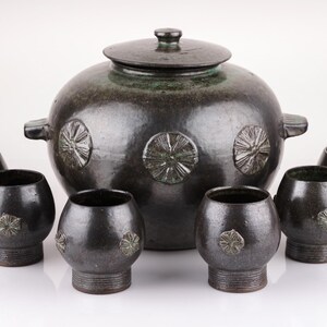 Könnte beinhalten: Ein Set aus sechs Keramik-Trinkgefäßen, darunter ein großer, mit Deckel versehener Krug mit rundem Körper und dekorativen Blumendesigns. Der Krug und die Becher sind alle dunkelbraun mit grünen Akzenten.