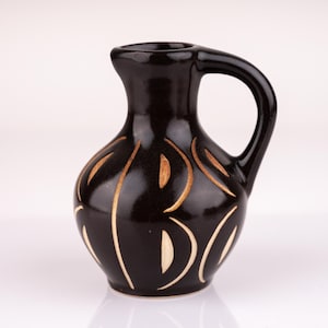 Könnte beinhalten: Ein kleiner, schwarzer Keramik Krug mit brauner Glasur und einem weißen Design. Der Krug hat einen runden Körper und einen kurzen, gebogenen Ausguss.