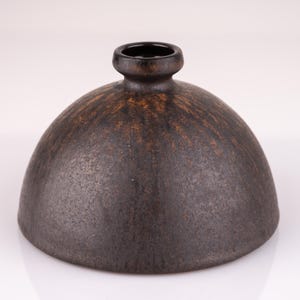 Puede incluir: Un jarrón de cerámica marrón oscuro con forma de cúpula redondeada y una abertura ligeramente acampanada. El jarrón tiene una superficie texturizada y un sutil glaseado marrón.