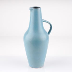 Könnte beinhalten: Ein hellblaues Keramikkännchen mit Henkel und schmalem Ausguss. Das Kännchen hat eine glatte, matte Oberfläche und einen leicht abgerundeten Körper. Die Innenseite des Ausgusses ist dunkel. Das Kännchen ist ein dekorativer Gegenstand.