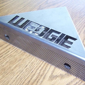 May include: A silver triangular metal corner guard with a perforated design and the word "WEDGIE" cut out in the center.
