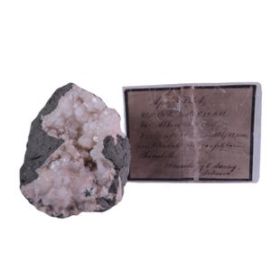 Puede incluir: Un espécimen geológico con un grupo de cristales de color rosa pálido incrustados en una matriz de roca gris oscura. Una etiqueta manuscrita con texto descolorido acompaña al espécimen, proporcionando detalles sobre su origen y composición.
