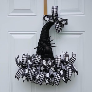 May include: A black Halloween witch hat wreath with a large spider and decorative ribbons. The ribbons feature a black and white checkered pattern and white ghosts with the word "BOO".
