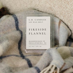 May include: A clear plastic clamshell container of C.B. Candles soy wax melt with a white label reading Fireside Flannel, Handpoured in Phillipsburg, NJ, and 2.7 oz | 76.5 g. The wax melt is positioned on a light blue and white plaid blanket.