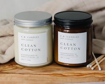 May include: Two soy candles in glass jars with silver and black lids. The candles are labeled "C.B. Candles Soy Candle Clean Cotton" and are hand-poured in Phillipsburg, NJ. The candles are sitting on a wooden surface.
