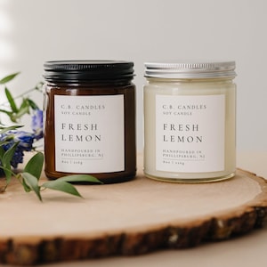 May include: Two soy candles in glass jars with black and silver lids. Both candles are labeled "C.B. Candles Soy Candle Fresh Lemon" and "Handpoured in Phillipsburg, NJ". The candle on the left has a brown jar and the candle on the right has a clear jar. Both candles are sitting on a wooden slice.