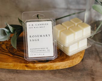 May include: A clear plastic container with six white wax melts labeled "Rosemary Sage" soy wax melt. The container is on a wooden surface.