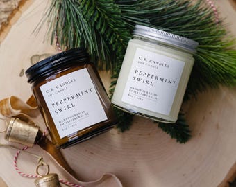 May include: Two glass jars of soy candles with black and silver lids, labeled "Peppermint Swirl." The jars are on a wooden surface with greenery and bells. Each candle is 8 oz / 226g.