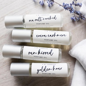 May include: Four small glass bottles with silver roll-on tops and white labels. The labels have black text that reads "C.B. CANDLES", "sea salt + orchid", "PERFUME OIL", "INGREDIENTS:", "cocoa cashmere", "sun kissed", "golden hour".