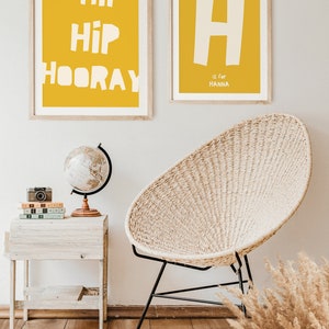 May include: Two framed prints on a white wall. The print on the left has a yellow background with the text "Hip Hip Hooray" in white. The print on the right has a yellow background with a white letter "H" and the text "is for Hanna" in white.