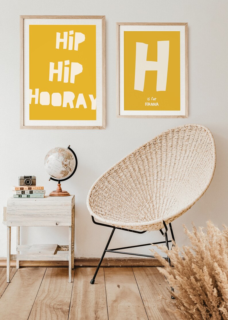 May include: Two framed prints on a white wall. The print on the left has a yellow background with the text "Hip Hip Hooray" in white. The print on the right has a yellow background with a white letter "H" and the text "is for Hanna" in white.