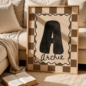 Puede incluir: Impresión artística enmarcada con una gran letra negra "A" y el nombre "Archie" en cursiva. La obra de arte tiene un fondo beige y un borde a cuadros marrón y beige. La impresión está enmarcada en un marco de madera.