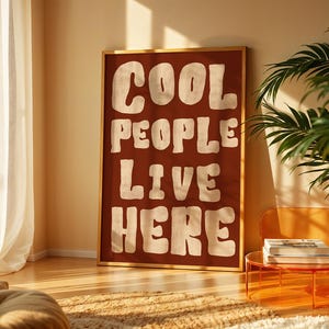 May include: A framed art print with the words "COOL PEOPLE LIVE HERE" in large, white, retro-style lettering on a dark red background. The print is in a gold frame and leans against a wall in a room with a plant and a small table.