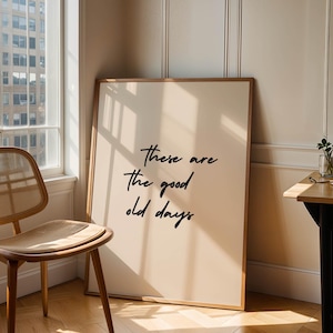 May include: A framed art print with the text "these are the good old days" in black script. The print leans against a wall next to a wooden chair and a window. The room is bathed in natural light.