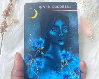 Könnte beinhalten: Eine Tarotkarte mit blauem Hintergrund und einem Halbmond in der oberen linken Ecke. Die Karte zeigt eine Frau mit blauer Haut und blauen Blumen. Der Text "INNER GODDESS" ist in Gold oben auf der Karte gedruckt.