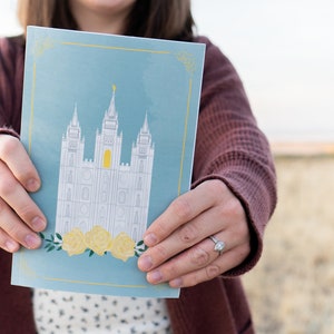 Puede incluir: Una tarjeta azul con una ilustración blanca de un templo con acentos amarillos y un diseño floral en la parte inferior. La tarjeta dice "Temple"
