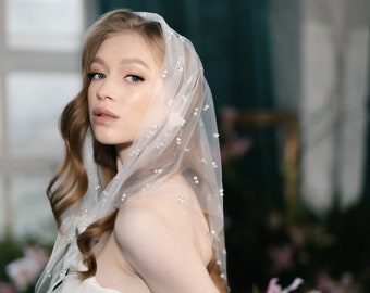 May include: A bride wearing a white satin wedding dress and a sheer veil adorned with small pearls. The veil covers the head and shoulders, and the sleeves are sheer with pearl accents. The background features flowers and a window.