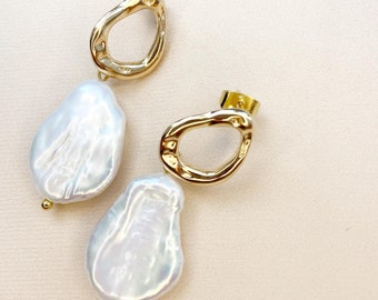 May include: A pair of gold-colored, open-circle stud earrings with large, irregular, pearlescent white baroque pearl drops. The earrings are displayed on a light beige surface.