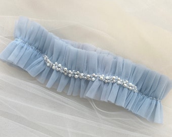 May include: A light blue garter with a row of pearl-like embellishments. The garter is made of tulle and has a ruffled edge.