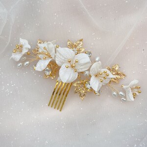 Op de afbeelding: Een goudkleurige haar kam met witte bloemaccenten, parels en heldere kristallen. De kam heeft gouden tanden en is versierd met gedetailleerde gouden bladeren. Het ontwerp is elegant en geschikt voor bruiloften of formele gelegenheden.