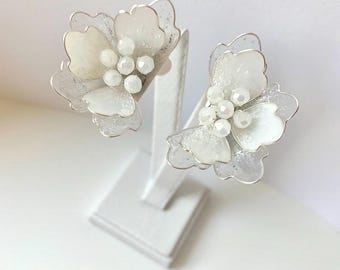 May include: A pair of flower-shaped earrings featuring translucent white petals with silver wire edges and a cluster of small, round, white beads at the center. The petals have a subtle, shimmering, glittery texture throughout their surface.