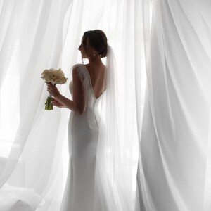 May include: A bride in a white wedding dress with a long veil, holding a bouquet of white flowers, stands in front of a white sheer curtain.