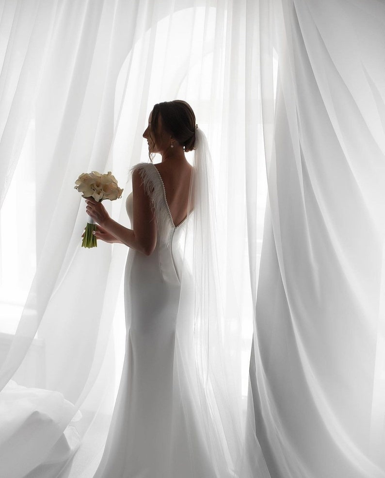 May include: A bride in a white wedding dress with a long veil, holding a bouquet of white flowers, stands in front of a white sheer curtain.