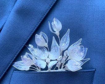 May include: A delicate, clear floral lapel pin with white and light purple accents, set in the pocket of a navy blue blazer. The pin features multiple flower buds and leaves, creating an elegant accessory.