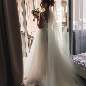 May include: A bride in a white wedding dress with a long veil and a bouquet of white flowers stands by a window.