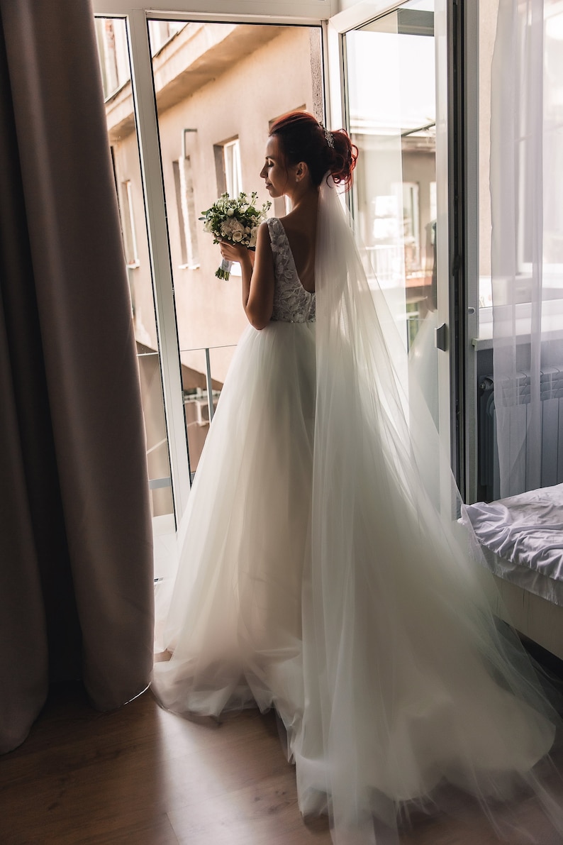 May include: A bride in a white wedding dress with a long veil and a bouquet of white flowers stands by a window.