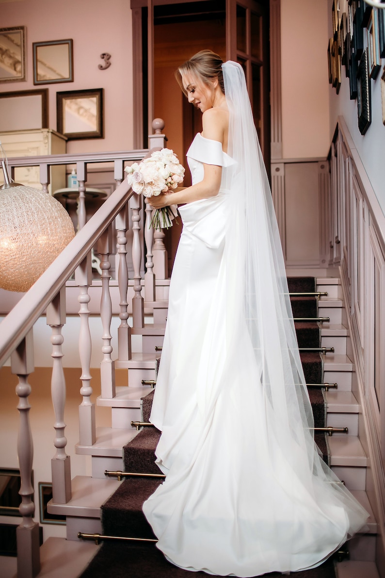 May include: A bride in a white wedding dress and veil walks down a staircase with a bouquet of white flowers.