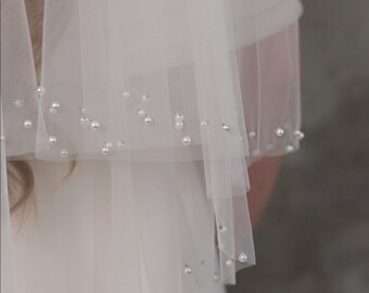 May include: A sheer white bridal veil with multiple layers, adorned with scattered faux pearls. The veil drapes over a white dress, creating a soft, elegant look. The pearls add a touch of sparkle to the delicate fabric.