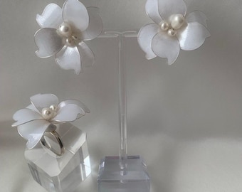 May include: A set of floral jewelry, including earrings and a ring. The earrings and ring feature white, petal-shaped designs with a pearl center. The earrings are displayed on a clear stand, and the ring rests on a clear cube.