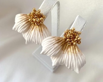 May include: A pair of white flower earrings with gold accents. The earrings feature a layered petal design with gold detailing on the edges and a cluster of gold beads at the center, displayed on white stands.