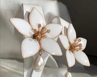 May include: A pair of white flower earrings with gold-colored accents. Each earring features a cluster of gold-colored beads at the center and a dangling pear-shaped pearl. The petals have a gold outline.