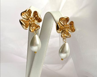 May include: A pair of gold-toned earrings featuring a four-petaled flower stud design with a textured center. A smooth, white, teardrop-shaped faux pearl hangs from each flower, secured by a small gold-toned bead at the base of the pearl.
