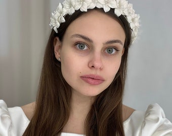 May include: A white floral headband featuring a series of fabric flowers arranged in a crown formation. Each flower is accented with small, clear, translucent beads at the center, creating a textured and detailed appearance across the entire band.