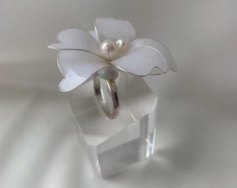 May include: A silver ring featuring a white flower design with a pearl center. The flower petals are white with silver edges. The ring is displayed on a clear, rectangular stand.