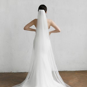 Puede incluir: Un vestido de novia blanco con una larga cola y un velo transparente. El velo est&aacute; sujeto a la parte posterior de la cabeza y cae sobre el vestido. El vestido es sencillo y elegante.