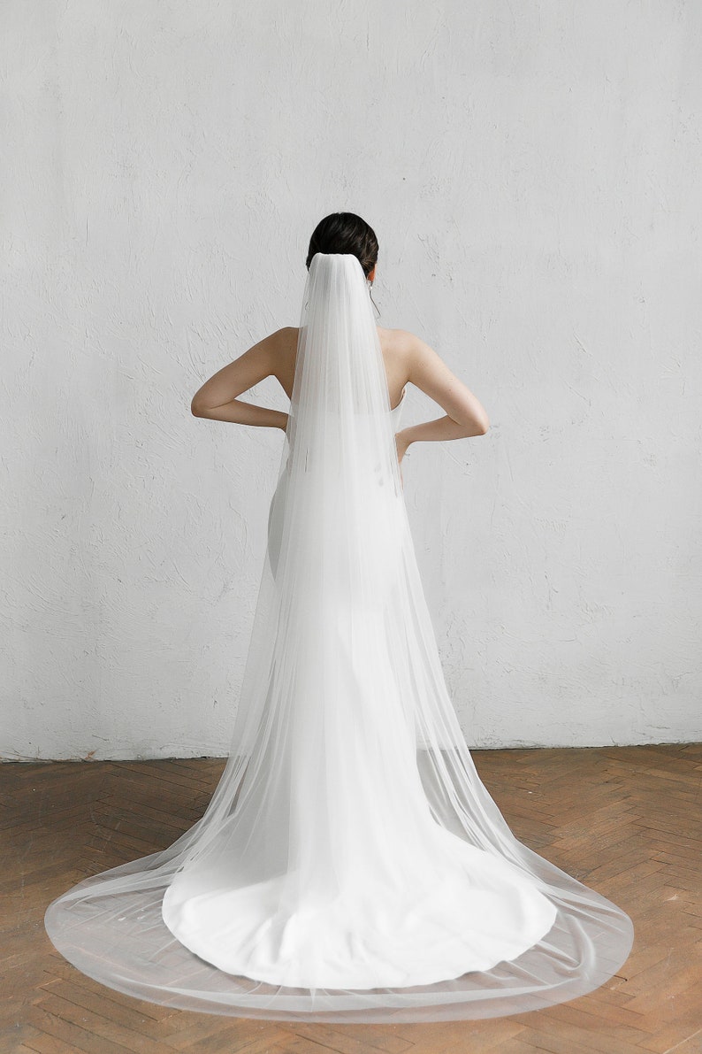 Puede incluir: Un vestido de novia blanco con una larga cola y un velo transparente. El velo est&aacute; sujeto a la parte posterior de la cabeza y cae sobre el vestido. El vestido es sencillo y elegante.