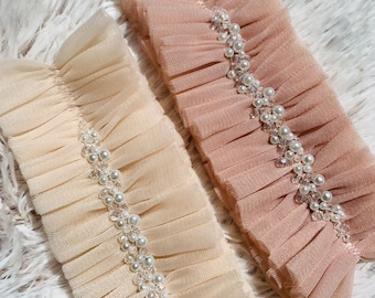 May include: Two ruffled tulle accessories, one in a soft peach and the other in a cream color. Both are adorned with a delicate arrangement of faux pearls and clear beads. The accessories are likely for a special occasion.