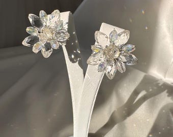 May include: A pair of clear crystal flower earrings. Each earring features a central cluster of crystals surrounded by larger, petal-shaped crystals. The earrings are displayed on a white stand against a soft, white background.