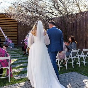 May include: A bride in a white wedding dress and veil walks down the aisle with her father. Guests are seated in white chairs, watching the ceremony.